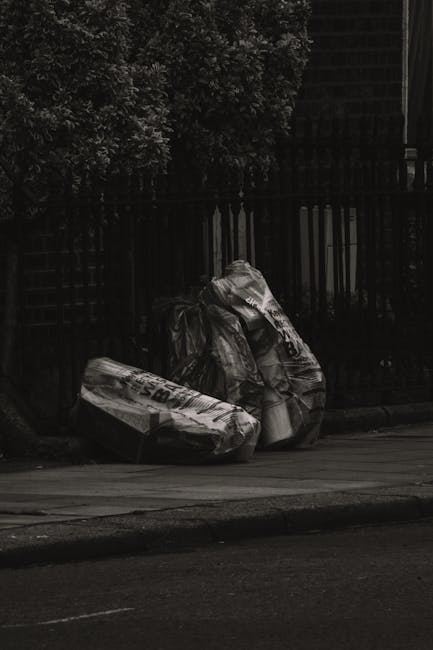 Two large black plastic rubbish bags, filled and tied at the top, are placed on a pavement next to a low curb in an outdoor urban setting. The bags appear to contain mixed waste materials, with one showing some printed text on its surface. They are positioned in front of a black metal fence with vertical bars, behind which dense foliage of a tree is visible at the upper left corner of the image. The scene is lit with natural light, casting subtle shadows on the pavement and emphasizing the texture of the bags and the nearby road surface, which has a slight incline. The environment suggests a typical residential or street-side area where waste has been temporarily placed for collection, indicative of private waste handling or independent rubbish removal services such as those provided by companies like Waste Removal Brompton. The overall atmosphere is neutral and functional, fitting with the context of rubbish clearance or waste disposal services aimed at maintaining cleanliness in urban outdoor spaces.