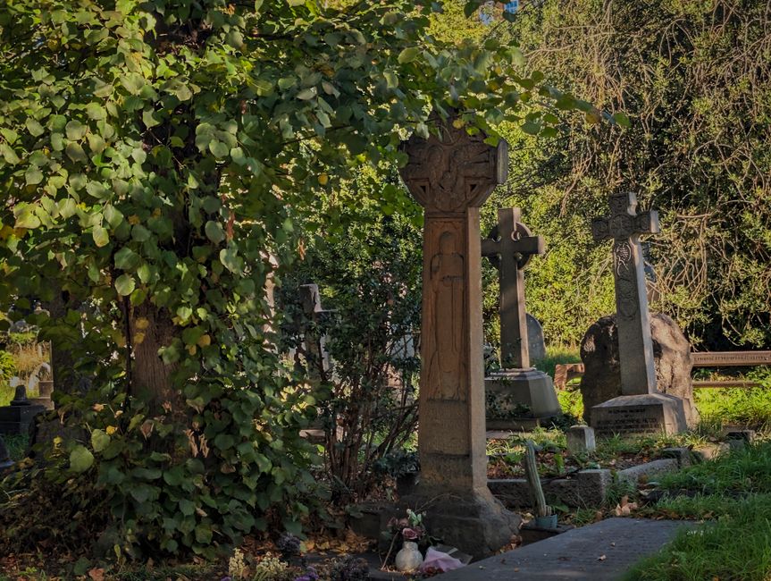 A cemetery scene featuring several traditional stone gravestones with carved crosses and ornamental details, surrounded by lush green foliage and mature trees. The central gravestone is tall and narrow with intricate carvings, while other stones in the background vary in size and shape, some rounded or with decorative tops. The gravestones are weathered, with some moss or lichen growth visible on their surfaces, indicating age. The ground is uneven and grassy with some small flower arrangements and lighting candles placed near a few markers. Dense shrubbery and leaves partially obscure parts of the graves, creating a tranquil, natural environment. Natural sunlight filters through the foliage, casting soft shadows across the scene. This quiet burial site is typical of an older cemetery that relies on traditional stone monuments, and it reflects a setting where private or independent waste collection services, such as those provided by Waste Removal Brompton, might be involved in clearing or maintenance activities, as suggested by the context of rubbish removal services in the area.