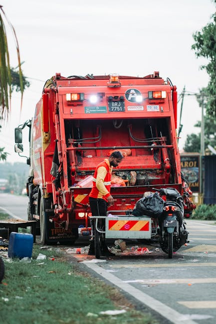 A waste collection worker is seen operating a large red rubbish truck parked on the side of a street. The worker, dressed in a high-visibility orange vest over a dark shirt and trousers, is actively working at the rear of the vehicle, which has an open compartment with mechanical arms for lifting and collecting waste. The truck displays various reflective decals and identification labels, including a number plate reading 'BRJ 7751', and a small distance indicator sign. In front of the truck, a black motorcycle with a large black plastic bag on its seat is parked, partially obstructed by a temporary barrier with reflective red and white stripes. The surrounding environment includes a road with a marked curb, an adjacent grassy verge, and some trees and structures in the background, with an overcast sky. This scene illustrates a typical instance of private waste collection or rubbish removal service, akin to that provided by companies like Waste Removal Brompton, in an urban or suburban setting.