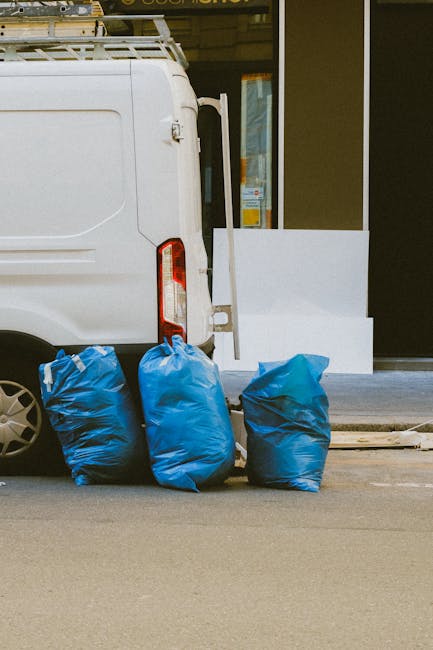 The image depicts the rear side of a white commercial van parked on a paved street in front of a building with large glass doors. Three large blue plastic rubbish bags, filled with waste, are placed on the ground close to the van's rear, aligned against its rear wheel arch. The bags are tightly gathered at the top, showing creases and a slight sheen on their surface, indicative of thick, durable plastic material. The van's rear door and taillight are visible, with the taillight featuring a vertical red and white design. In the background, the building's exterior wall is partially visible, with a smooth dark surface and a section of the storefront window. The scene is well-lit, suggesting daylight, and the setting appears to relate to on-site rubbish collection or private waste disposal, possibly facilitated by Waste Removal Brompton as part of their rubbish removal services.