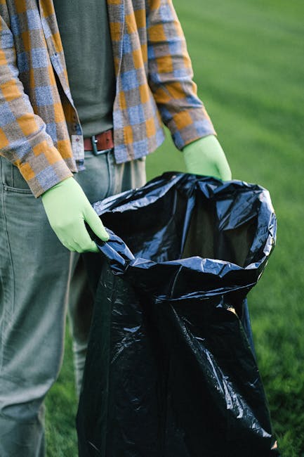 A waste collection worker is seen operating a large red rubbish truck parked on the side of a street. The worker, dressed in a high-visibility orange vest over a dark shirt and trousers, is actively working at the rear of the vehicle, which has an open compartment with mechanical arms for lifting and collecting waste. The truck displays various reflective decals and identification labels, including a number plate reading 'BRJ 7751', and a small distance indicator sign. In front of the truck, a black motorcycle with a large black plastic bag on its seat is parked, partially obstructed by a temporary barrier with reflective red and white stripes. The surrounding environment includes a road with a marked curb, an adjacent grassy verge, and some trees and structures in the background, with an overcast sky. This scene illustrates a typical instance of private waste collection or rubbish removal service, akin to that provided by companies like Waste Removal Brompton, in an urban or suburban setting.