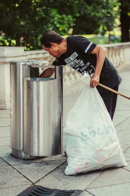 A middle-aged woman with short dark hair, wearing a black t-shirt with white text, is seen discarding a white garbage bag into a large, stainless steel public litter bin positioned on a paved sidewalk. The bin is cylindrical with an open top, set against a backdrop of lush green trees and a low stone or concrete balustrade. The woman is standing slightly bent forward, holding the bag with her left hand, while her right hand is partially obscured but appears to be holding a long wooden stick or litter picker. The scene is outdoors during daylight, with natural light illuminating the area and casting soft shadows. This image highlights an on-site waste disposal activity, aligning with private rubbish collection or street cleaning services, with the stainless steel bin indicating a maintained public space managed by services like Waste Removal Brompton. The setting suggests an organized effort for rubbish removal in a residential or park area, typical of alternative waste handling methods away from official council collections.