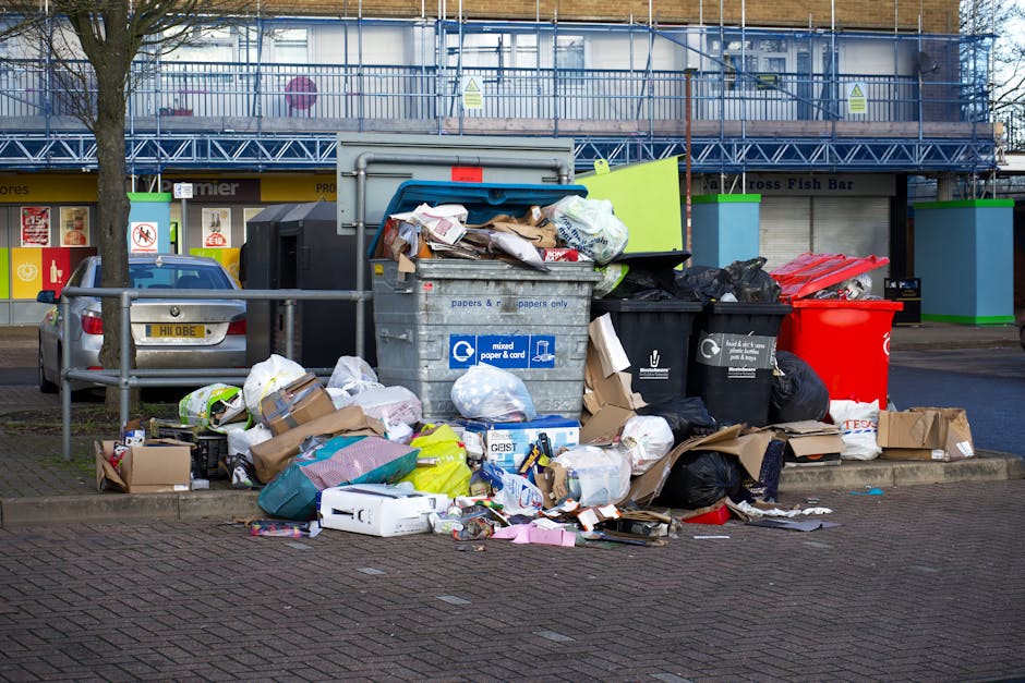 A large outdoor waste collection area featuring multiple overflowing rubbish bins, including a central metal bin labeled for mixed paper and cardboard, with its lid open and filled with various paper waste, cardboard boxes, and plastics. Surrounding the central bin are several black and red trash bags, some partially torn, and assorted discarded packaging, including flattened boxes, plastic containers, and wrapping materials scattered on the paved ground. To the left, a parked silver hatchback car is positioned behind a metal barrier, adjacent to a tree with bare branches. The background shows a commercial building with a blue metal scaffolding structure, a yellow storefront with signage indicating a fish bar, and closed shutter doors. The scene appears to be in an urban setting, highlighting concerns related to private waste disposal and rubbish removal services provided by Waste Removal Brompton, supporting alternative approaches to municipal waste collection.