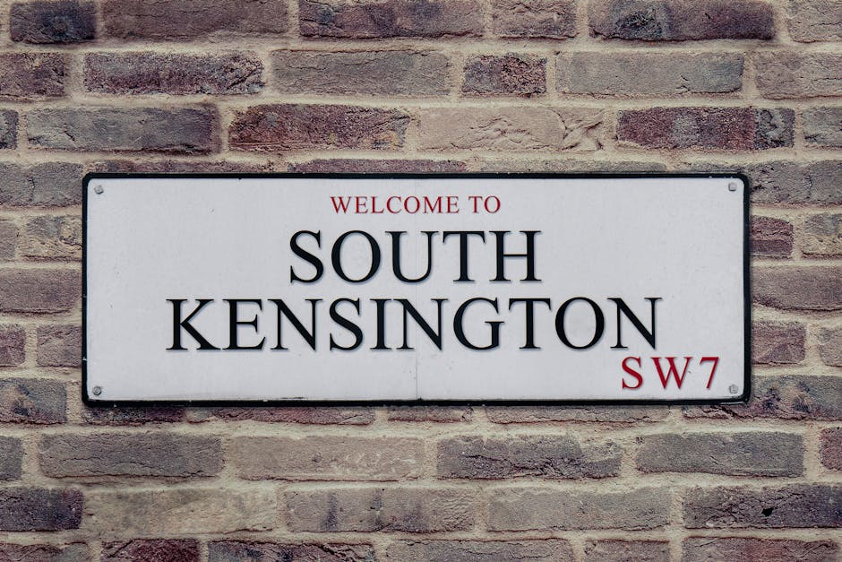A rectangular metal street sign mounted on a red-brown brick wall displaying the text 'Welcome to SOUTH KENSINGTON SW7' in black and red lettering. The sign has a white background with slightly rounded corners and appears to be secured with four screws, one at each corner. The brick wall behind the sign shows a traditional running bond pattern with textured, weathered bricks in shades of reddish-brown, light brown, and gray. The scene is evenly lit, without strong shadows or reflections, giving a clear view of the sign and the surrounding brickwork. This setting suggests a typical urban environment where private signage may be used for local identification or directional purposes, aligning with the context of waste removal and independent collection services by companies like Waste Removal Brompton operating in similar residential or commercial areas.