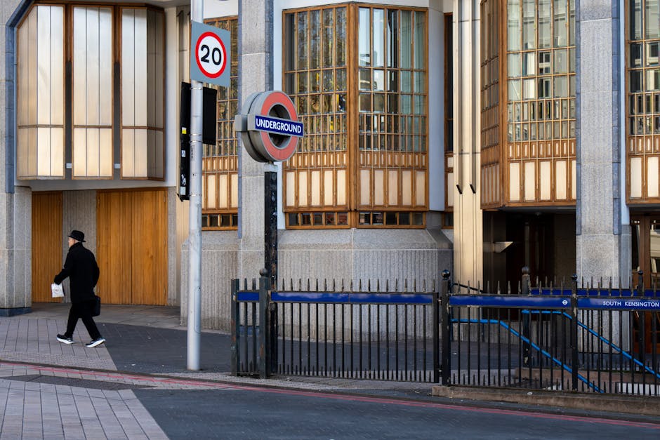 The image depicts a modern urban building with a facade featuring large, rectangular wooden-framed windows set into a concrete structure with vertical ridges. In the foreground, a black metal fence runs parallel to the pavement, with a section that appears to be an entrance or exit point. A pedestrian dressed in dark clothing, including a coat and hat, is walking along the sidewalk carrying a white plastic bag. Prominently displayed are two street signs: a circular speed limit sign indicating 20 miles per hour and an underground station sign with a red, white, and blue roundel. The scene is illuminated by natural daylight, highlighting the textures of the wooden window frames, the grey concrete, and the paving stones on the sidewalk, creating a neutral and professional urban environment that aligns with the theme of city waste and rubbish disposal, often associated with the need for efficient rubbish removal services near busy stations or commercial areas. The presence of the underground sign subtly emphasizes a location with independently managed waste collection options, consistent with private waste handling practices.