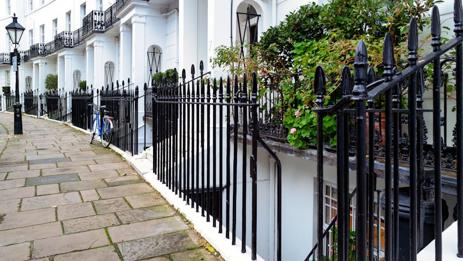 A row of white terraced buildings with black wrought iron railings along a paved sidewalk, featuring decorative finials on top of the fence posts. The pavement consists of large, rectangular stone slabs with visible joints, some with moss or dirt in the gaps. To the right, a garden area with lush green foliage and pink flowers is situated behind the iron fence, partially obscuring the lower part of the building and window beneath. A blue bicycle is leaned against the fence near the foreground, and a bollard is positioned along the sidewalk for pedestrian separation. The scene is lit naturally, indicating daytime, with soft shadows cast by the railing and foliage, creating a calm, orderly street environment that relates to private garden maintenance or outdoor clearance activities, possibly involving rubbish removal or garden waste management by Waste Removal Brompton in a typical residential setting.